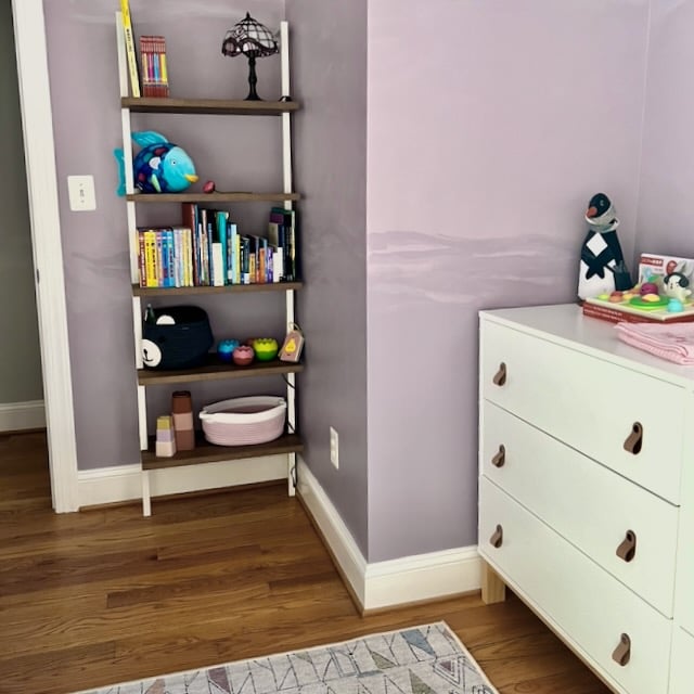 A ladder-style bookshelf filled with children’s books and toys, standing next to a white dresser with leather pulls in a room with light purple walls.