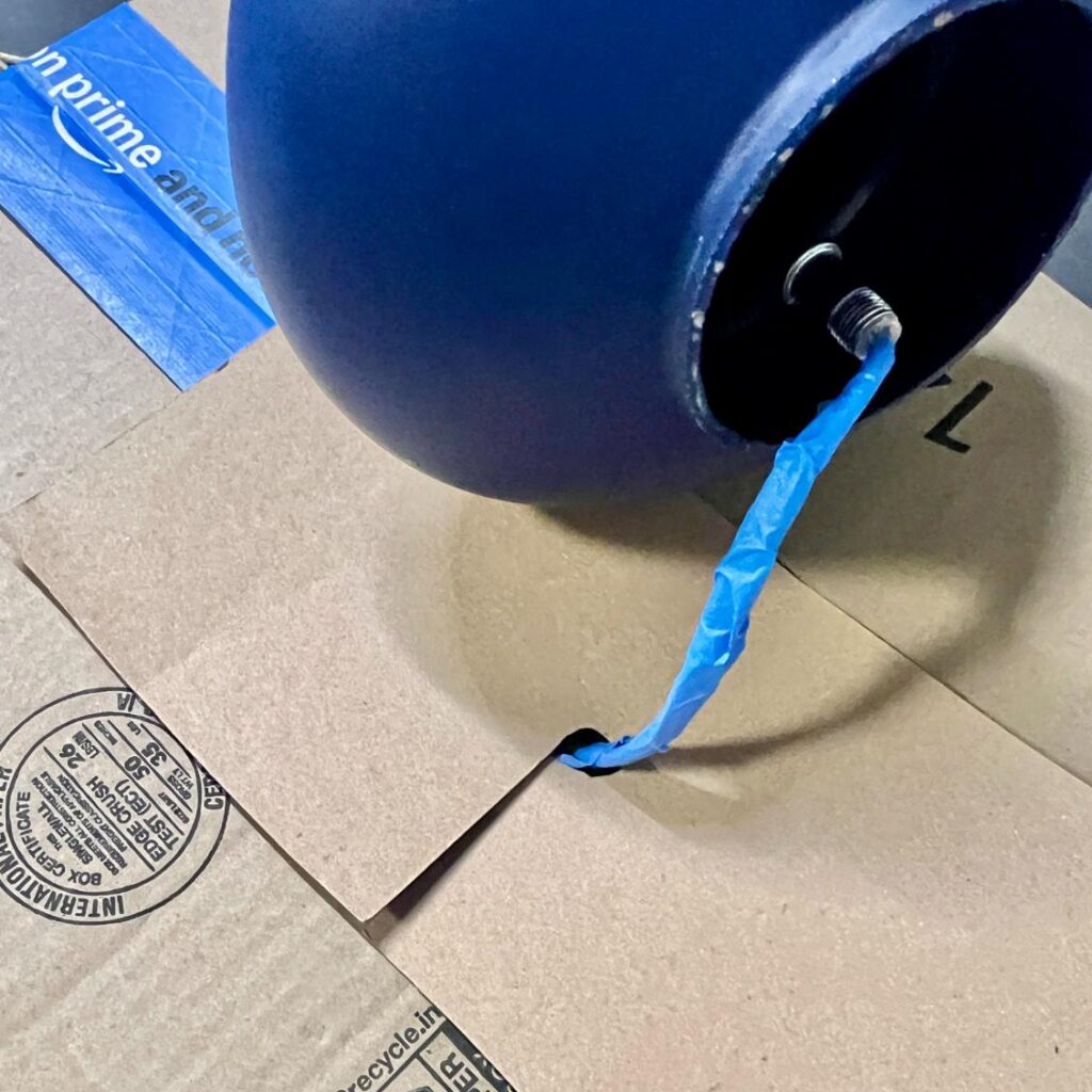 A blue, rope-wrapped, spherical pendant light fixture with a blue tape-covered wire, sitting on a cardboard shipping box.