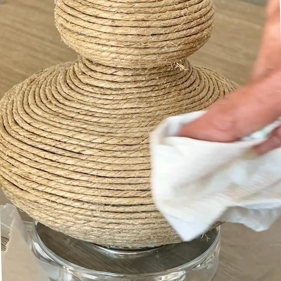 A hand wipes a rope-wrapped lamp base with a white cloth.