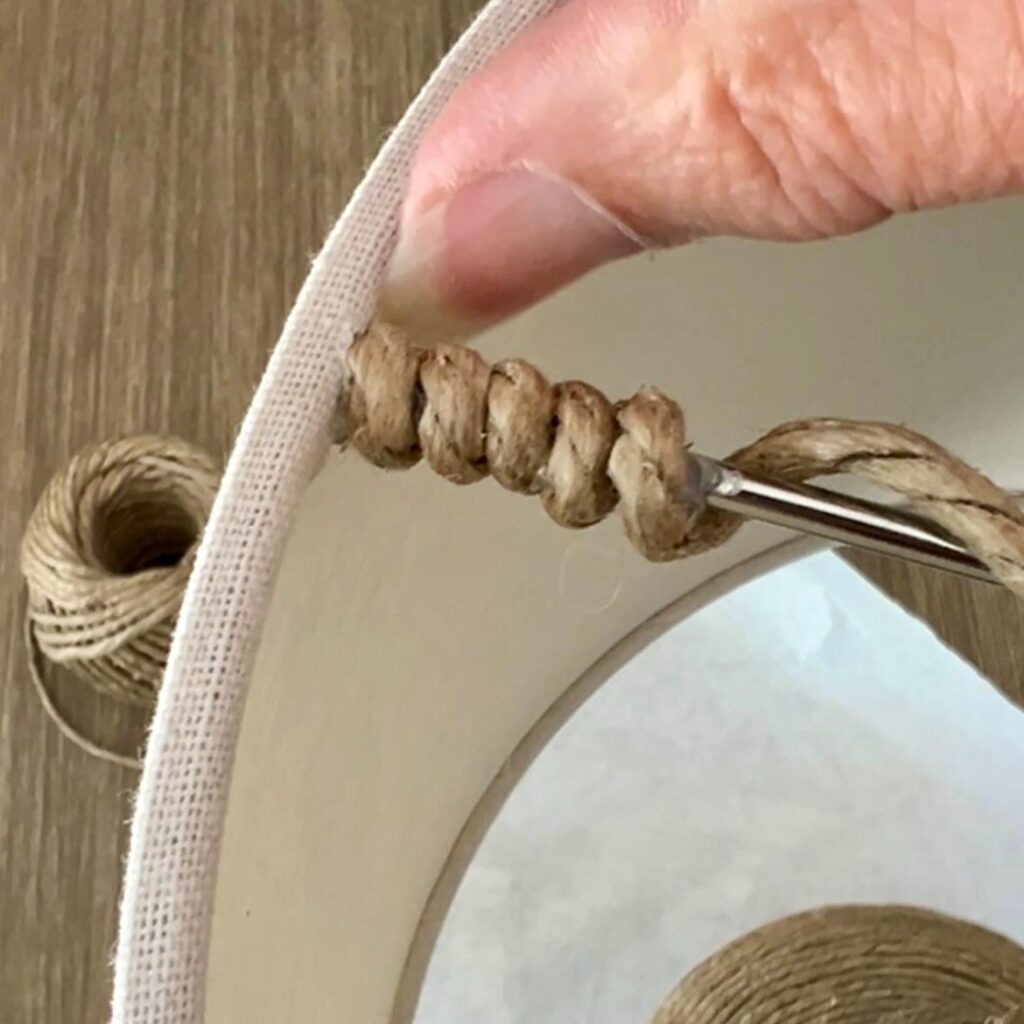 A person uses a crochet hook to pull a piece of twine through the fabric edge of a rope wrapped lamp.
