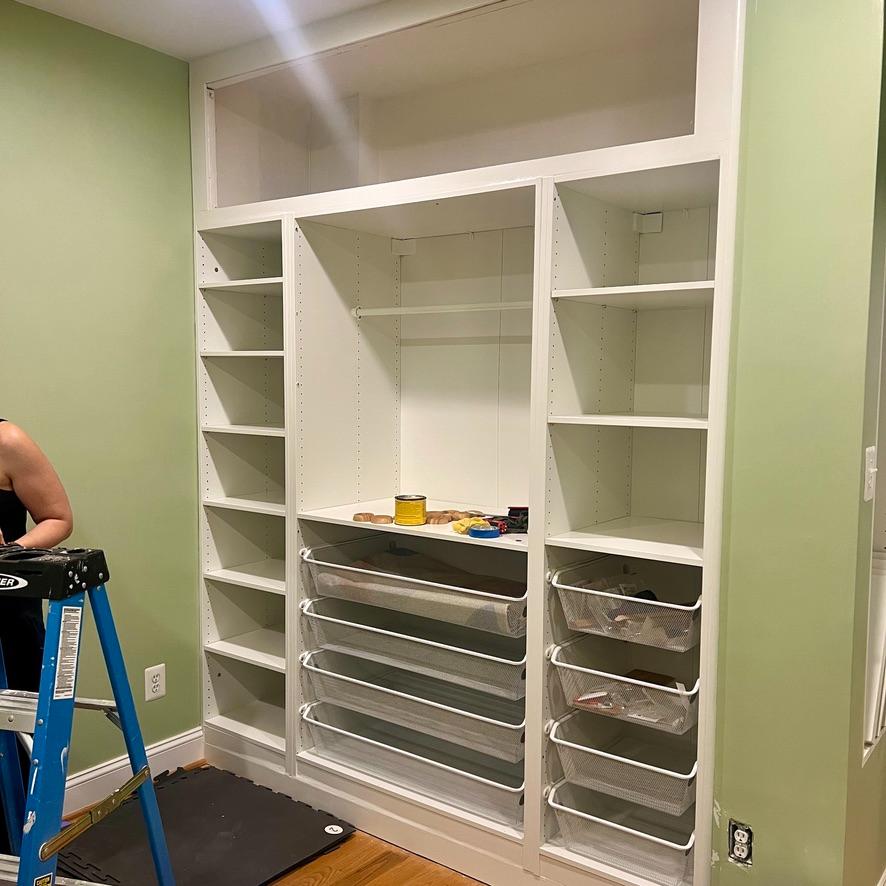 An IKEA Pax closet wall with open shelves, hanging space, and wire drawers installed against a light green wall.