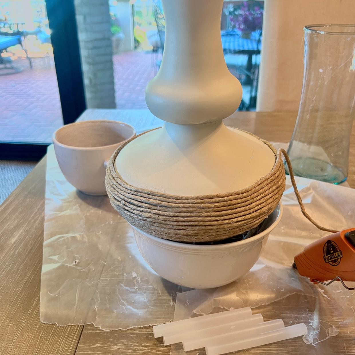 A white lamp base being wrapped with jute twine to create a rope wrapped lamp, with a glue gun and glue sticks nearby.