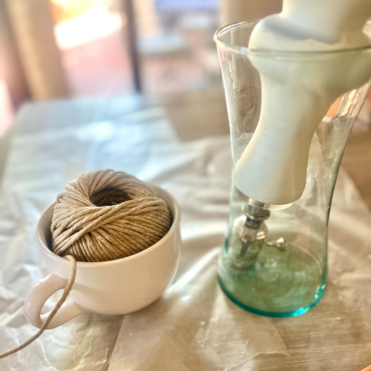 A ball of rope wrapped lamp twine in a white mug next to a glass vase containing a white object.