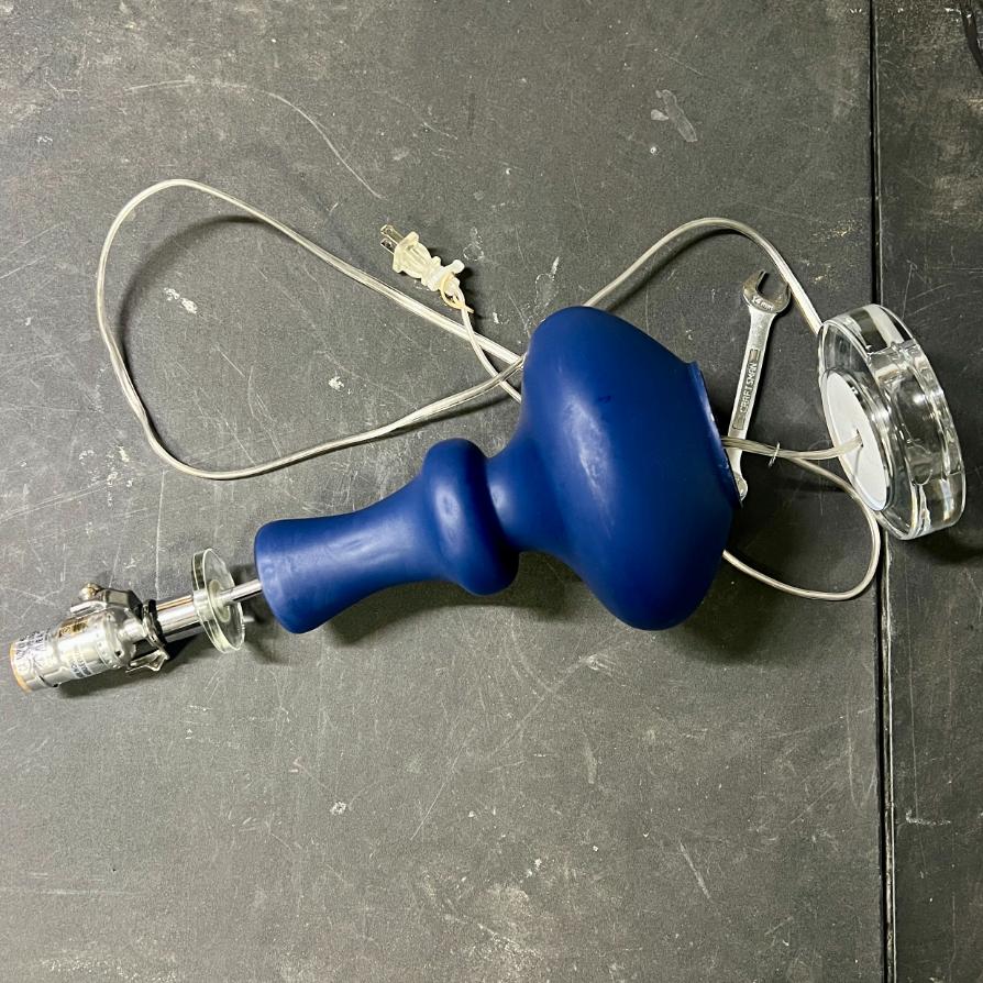 A blue rope wrapped table lamp with a curved base, a metal socket, and a transparent glass base plate, lying on a dark surface with a wrench hooked around its neck.