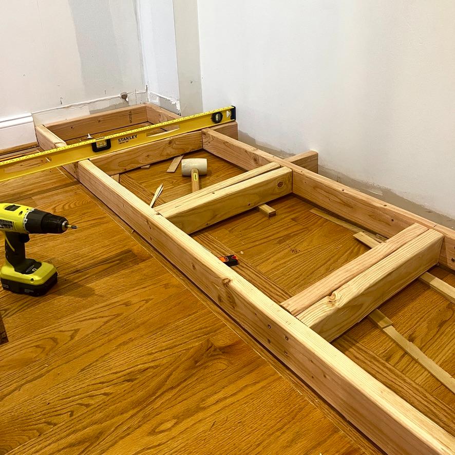 A wooden frame for an IKEA Pax closet wall under construction on a hardwood floor, with a level and a cordless drill nearby.