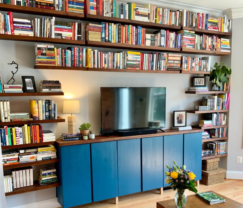 A wall featuring an IKEA Pax closet wall with doors, built-in wooden bookshelves, a mounted television, and a blue cabinet, with various books, a lamp, photos, and a potted plant displayed on the shelves.
