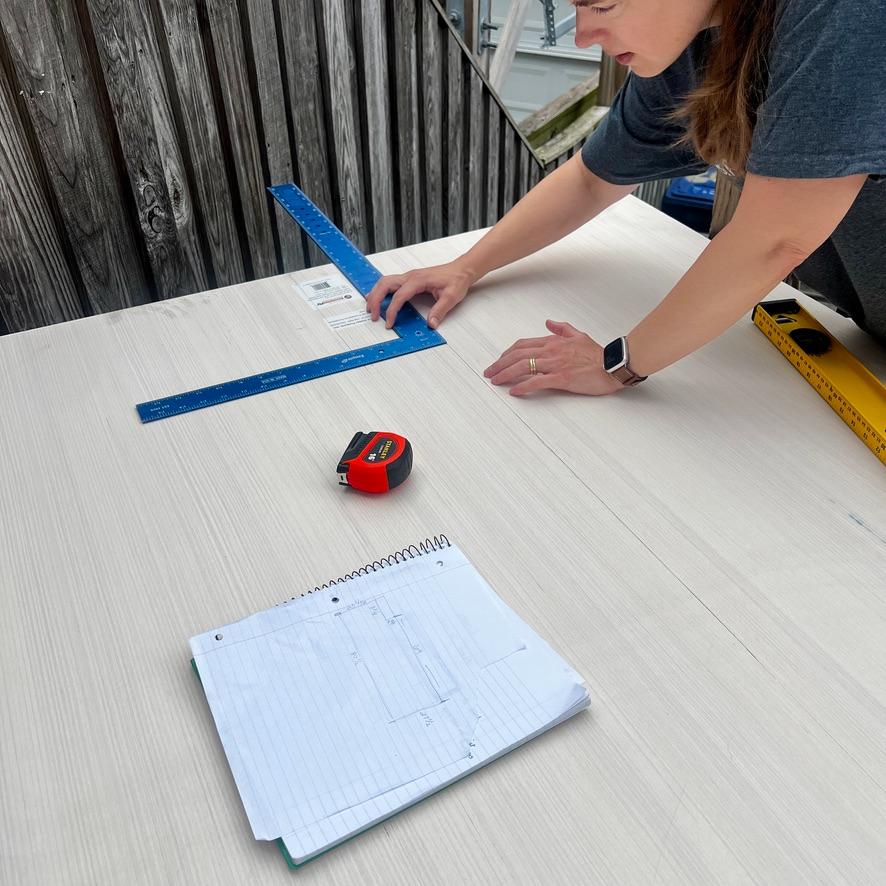 A person uses a blue carpenter’s square to mark a white table surface for an IKEA Pax closet wall, with a tape measure and a notebook containing a sketch nearby.