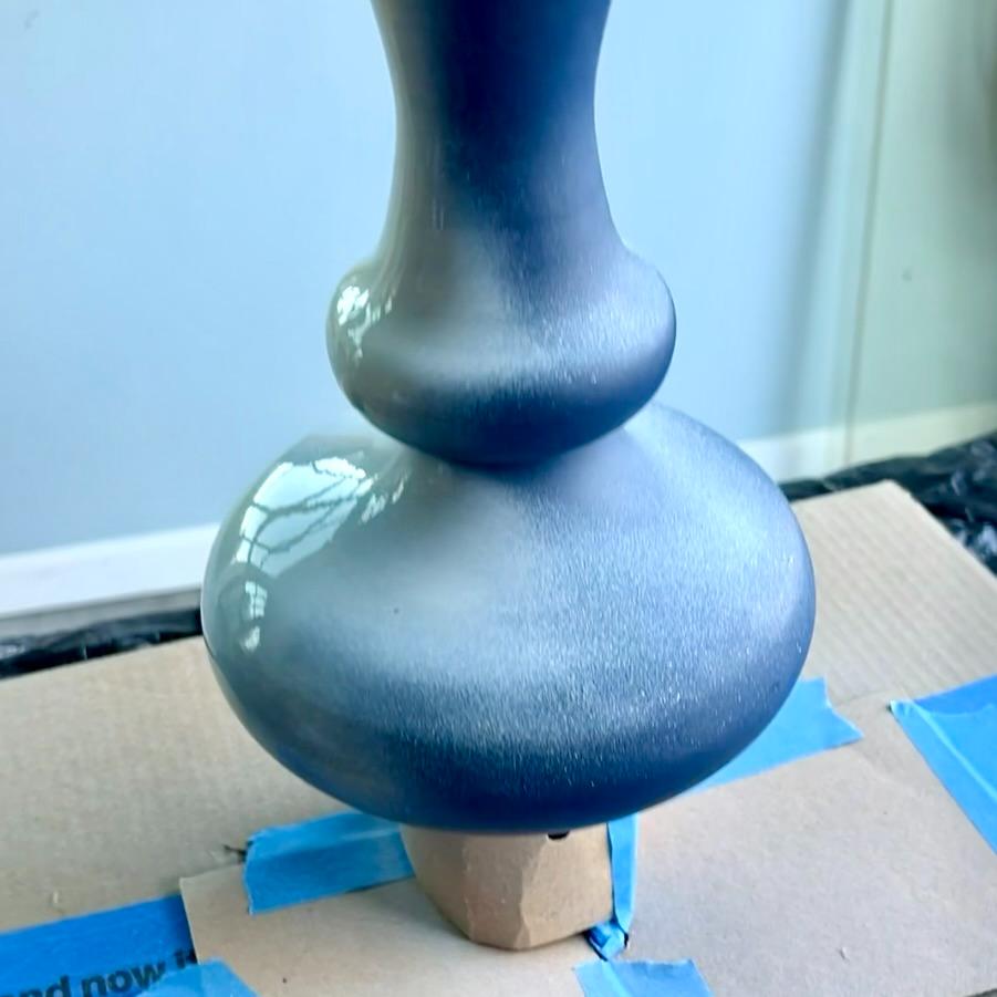 A bulbous, smooth gray vase that will become a rope wrapped lamp being spray-painted, set on a cardboard base with blue painter’s tape.
