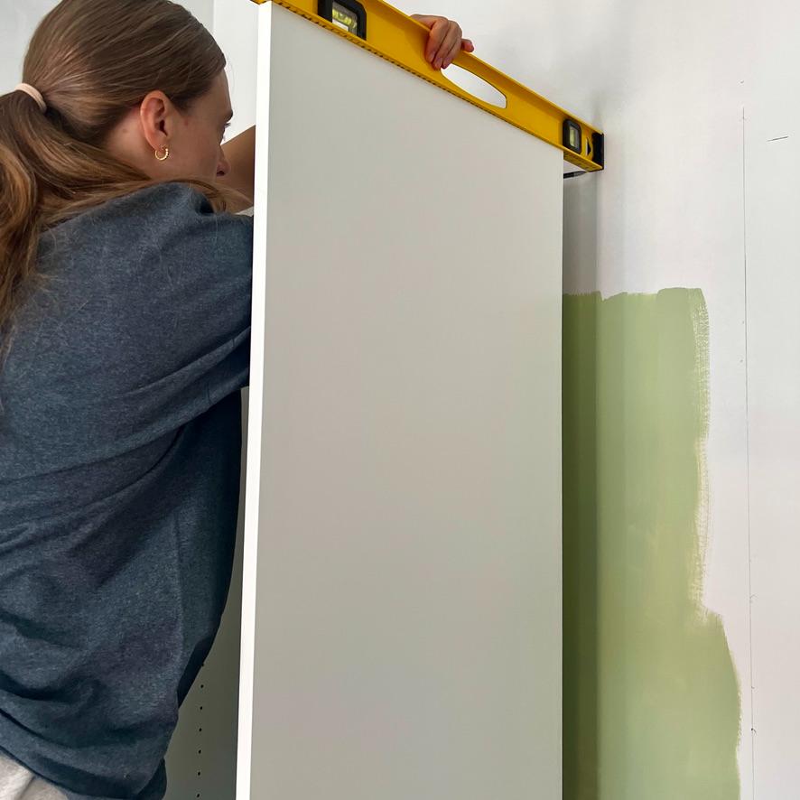 A person holds a yellow spirit level against the top edge of a white IKEA Pax closet door installed against a wall with a patch of green paint.