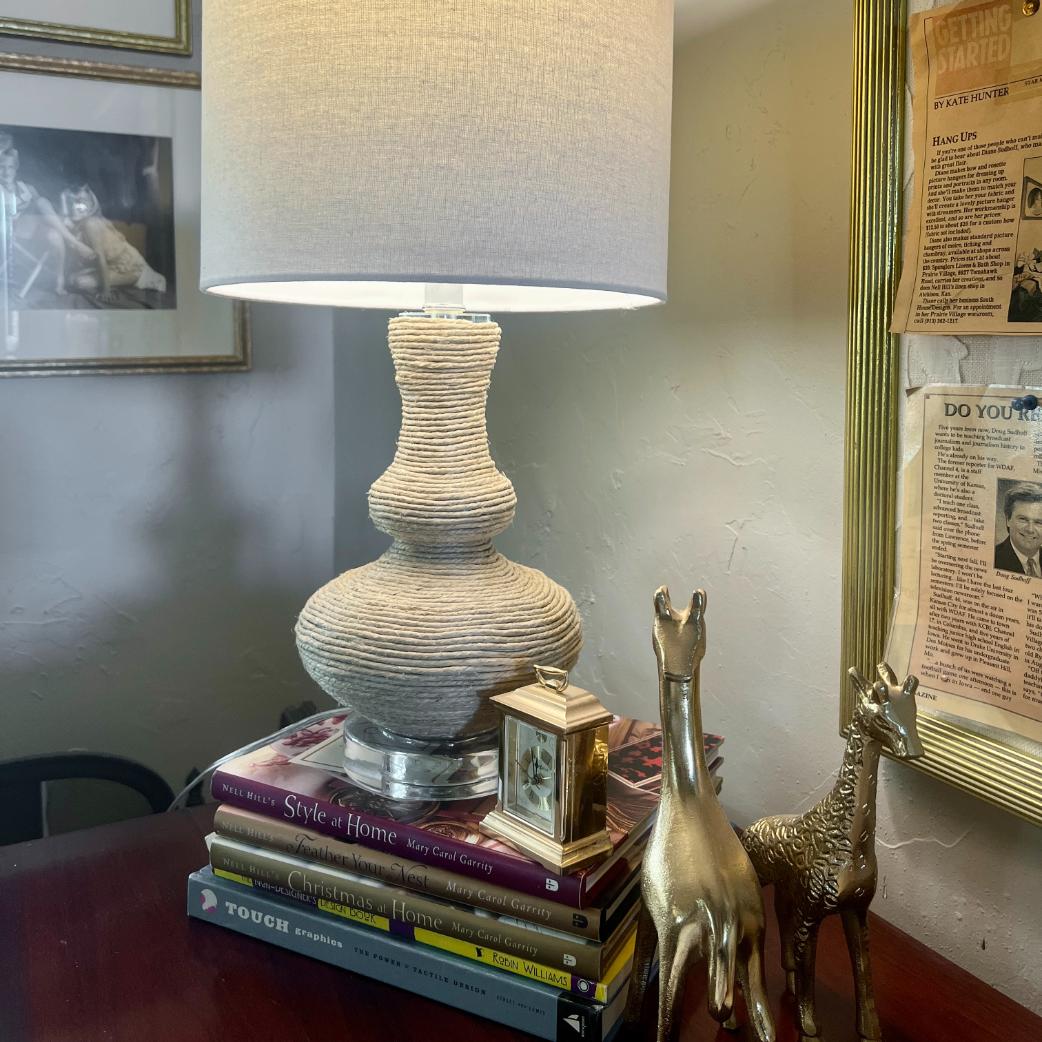 A rope wrapped table lamp with a ribbed white base sits on a stack of books, next to a small brass clock and two golden giraffe figurines.