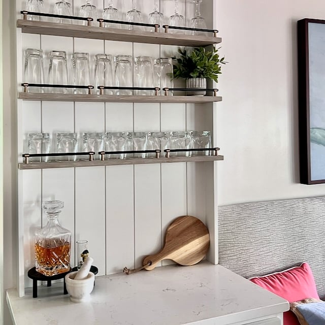 A white DIY storage shelving unit above pax wardrobes holds three rows of glassware, with a decanter of amber liquid, a mortar and pestle, and a small wooden board on the counter below.