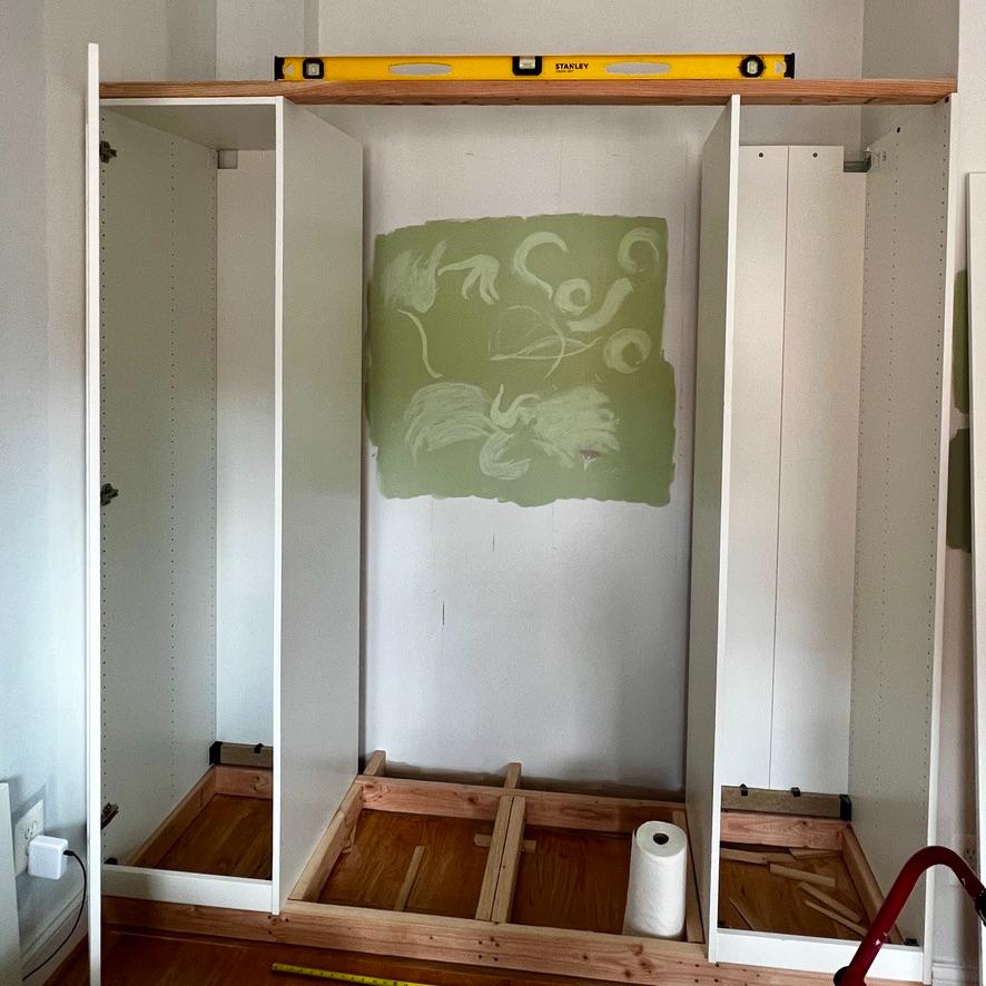 Two white IKEA Pax wardrobe frames installed on either side of an open space with wooden floor supports and a yellow level resting on top.