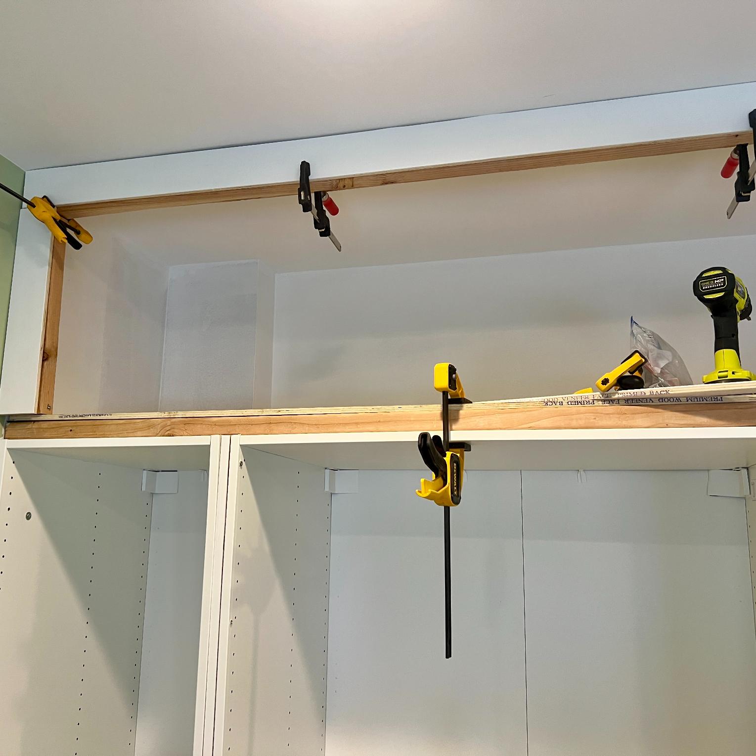 Wooden shelves for DIY storage above PAX wardrobes being installed over white cabinets, held in place by yellow and black clamps, with a cordless drill resting on the top shelf.