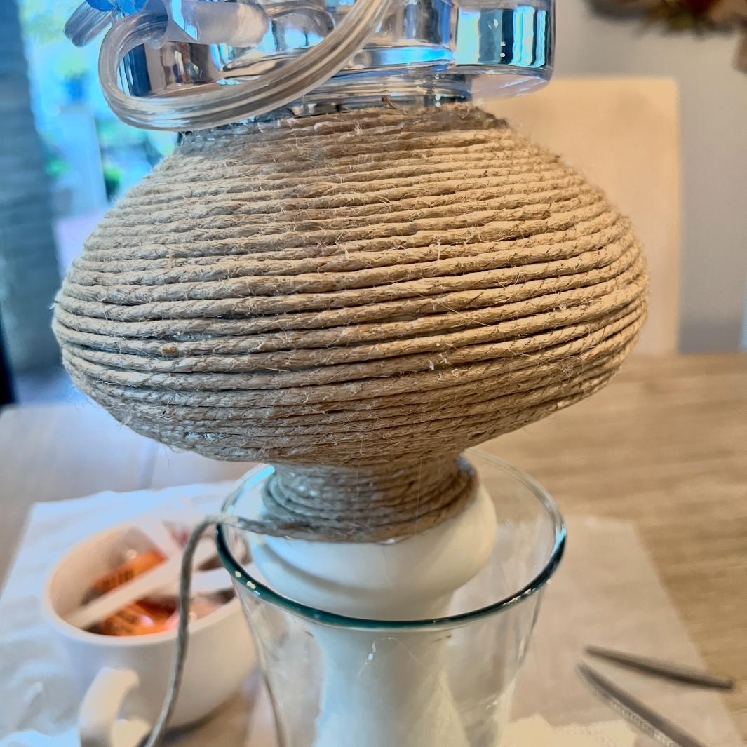 A glass vessel with a rope wrapped top section, finished in thick jute twine, sits on a wooden table.