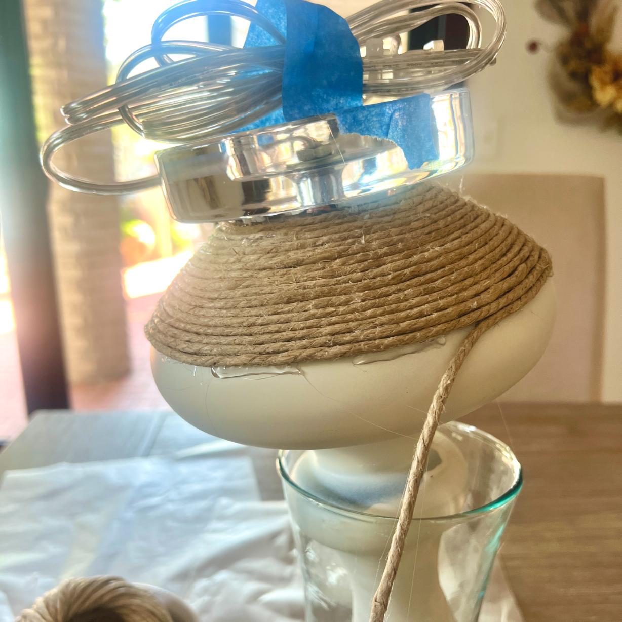 A rope-wrapped lamp base balanced on a glass and secured with blue painter’s tape.