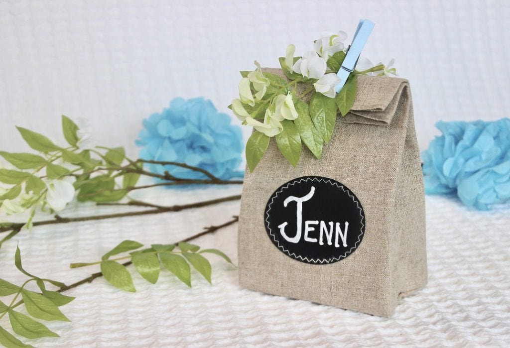 Linen party favor bag with an oval chalk cloth label sewn on that reads "Jenn". The bag is held closed with pretty blue clothespin and some silk green and flowers.
