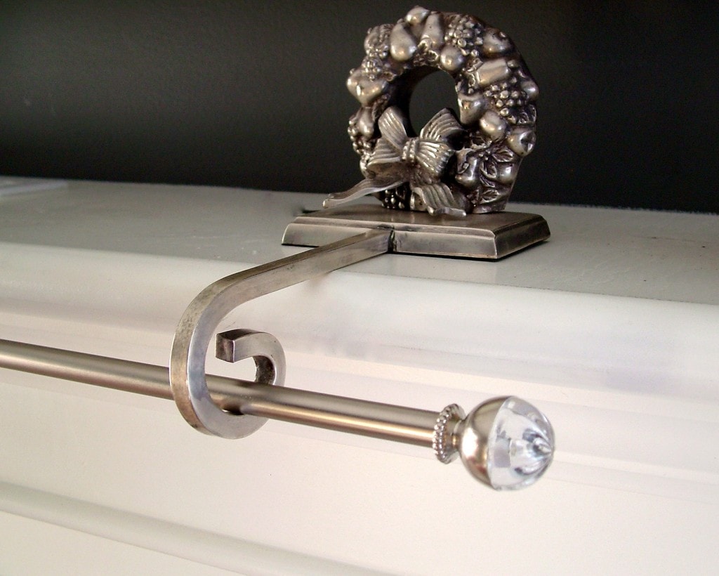 Silver wreath stocking holder suspending one end of a curtain rod on a white mantel with black wall.